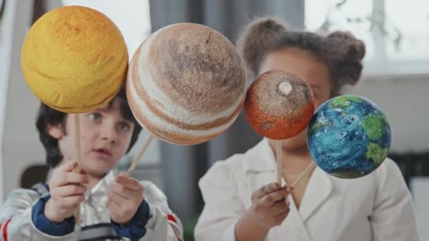 Lento tiro de inclinación hacia abajo de niño de 6 años en traje espacial y niña de 8 años en bata de laboratorio sosteniendo modelos de juguetes de planetas - Imágenes, Vídeo