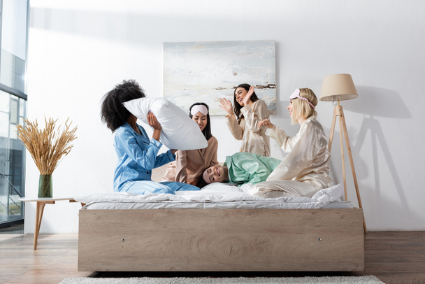 smiling interracial friends in pajamas having pillow fight during slumber party  - Foto, Imagen