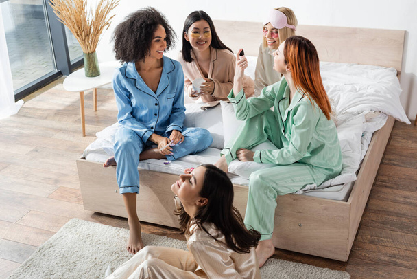 redhead woman showing nail polish to cheerful interracial friends during slumber party  - Photo, Image