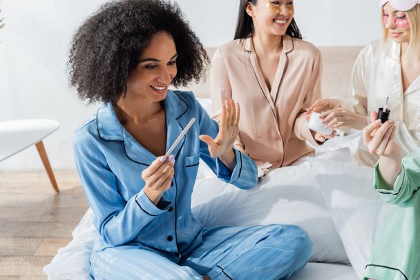 cheerful african american woman looking at nails while doing manicure near blurred friends during slumber party  - Photo, Image