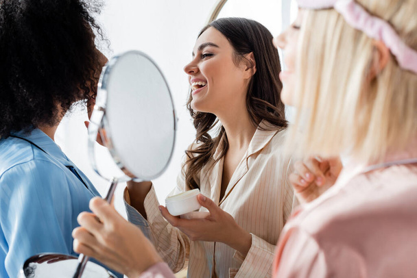 happy interracial friends applying face cream near blonde young woman with mirror on  blurred foreground  - Photo, Image