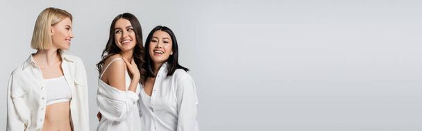 joyful multiethnic young models in white shirts smiling isolated on grey, banner - Foto, afbeelding