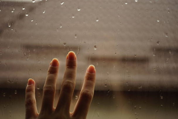 La mano toca una ventana con gotas de lluvia. Fondo del tejado en desenfoque. - Foto, imagen