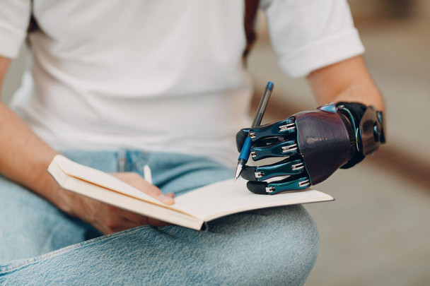 Young disabled man writes in notebook with pen in artificial prosthetic hand. - Photo, Image