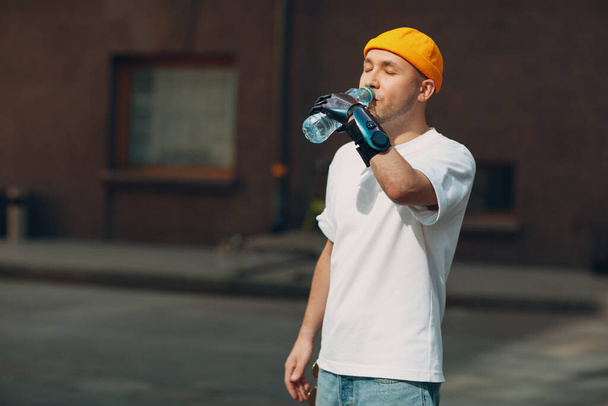 Νεαρός ανάπηρος άντρας κρατά και πίνει εμφιαλωμένο καθαρό νερό με τεχνητό προσθετικό χέρι σε casual ρούχα. - Φωτογραφία, εικόνα
