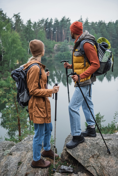 back view of couple with backpacks holding hiking sticks and looking at lake - Photo, Image