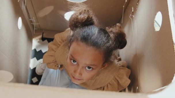 Toma de vista superior portátil de una linda niña de 8 años sentada dentro de un cohete de cartón y mirando a la cámara - Imágenes, Vídeo