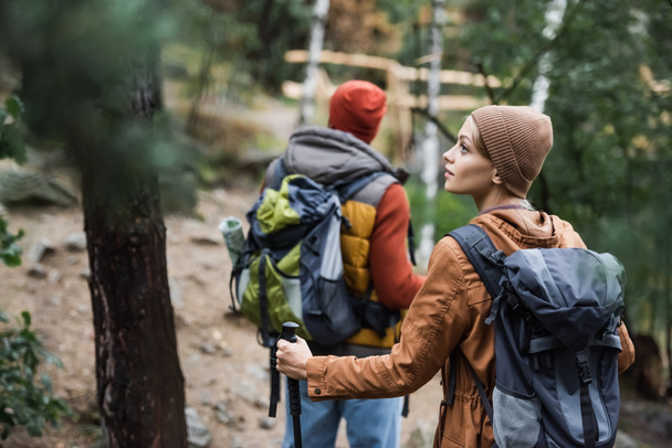 young woman with backpack looking away near man trekking in forest on blurred background - Foto, afbeelding
