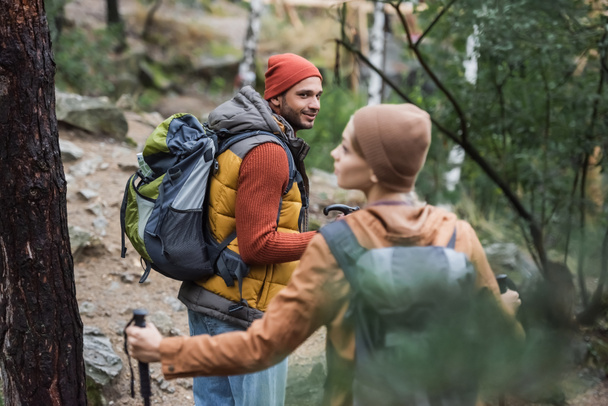 sonriente hombre con mochila mirando a la mujer trekking en el bosque en primer plano borroso  - Foto, imagen
