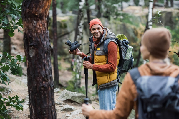 homme souriant tenant caméra vintage et regardant la femme trekking dans la forêt sur le premier plan flou  - Photo, image