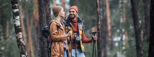 cheerful couple in hats holding hiking sticks while hugging in forest, banner - Фото, зображення
