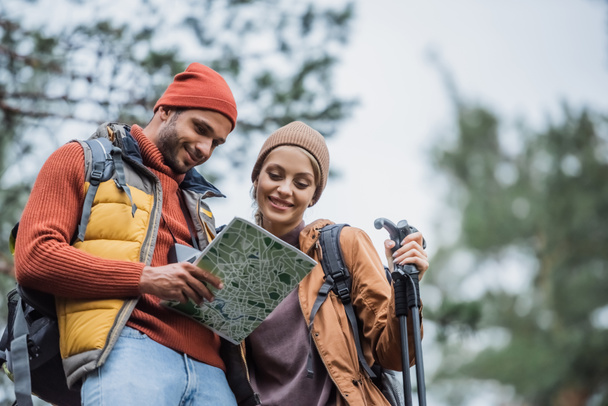 low angle view of happy couple in hats looking at map in forest - Foto, afbeelding