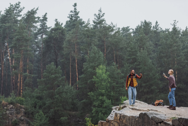 happy couple with smartphones standing on rocky cliff during rain - Photo, Image