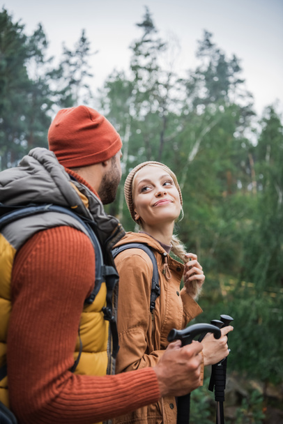 szczęśliwa młoda para z plecakami trekking razem z laski turystyczne i patrząc na siebie  - Zdjęcie, obraz