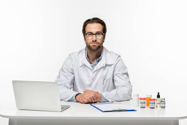 doctor en anteojos cerca de laptop, portapapeles y medicamentos medicinales de cannabis aislado en blanco - Foto, Imagen
