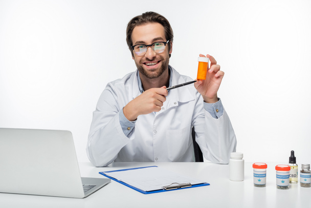 medico sorridente che punta con la penna al contenitore con pillole di cannabis medica isolate su bianco - Foto, immagini
