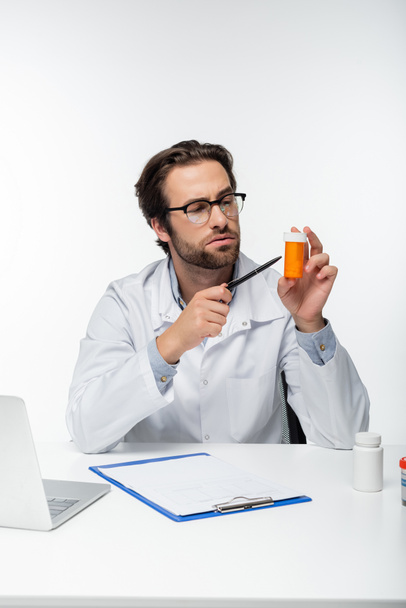 médecin pointant avec stylo au récipient avec des pilules de cannabis médical près du presse-papiers et ordinateur portable isolé sur blanc - Photo, image
