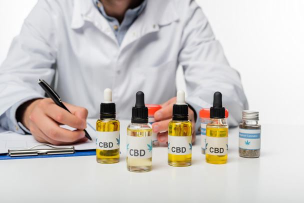 cropped view of doctor writing prescription near bottles with cbd oil isolated on white - Photo, Image