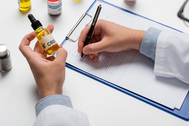 partial view of doctor with vial of cbd oil writing prescription on clipboard - Fotografie, Obrázek