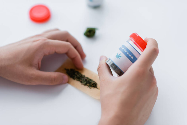 bijgesneden beeld van een man die een container met medicijnen vasthoudt terwijl hij een joint maakt met droge medische cannabis - Foto, afbeelding