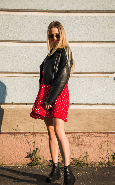 urban street portrait of young beautiful stylish woman in red dress and black leather jacket. Stands fooling around by the old blue wall - Фото, изображение