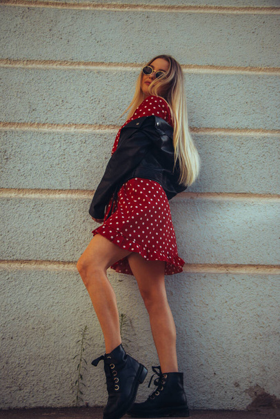 urban street portrait of young beautiful stylish woman in red dress and black leather jacket. Stands fooling around by the old blue wall - Φωτογραφία, εικόνα