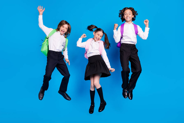 Photo of charming lucky schoolkids formalwear backpacks smiling jumping high rising fists isolated blue color background - Foto, afbeelding