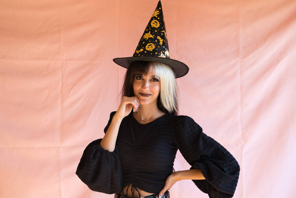 ブロンドと茶色の髪をした若い女性ハロウィーンパーティーの魔女の格好をして。彼女は魔女の帽子と黒のドレスを着ている. - 写真・画像