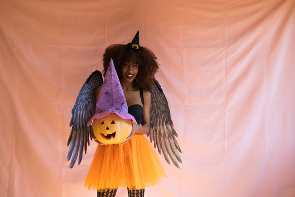 Νεαρή Αφροαμερικανή με στολή μάγισσας για το αποκριάτικο πάρτι. Φοράει καπέλο μάγισσας, πορτοκαλί φούστα και μαύρα φτερά αγγέλου. Η γυναίκα χαμογελάει στην κάμερα ενώ παίζει με την κολοκύθα. - Φωτογραφία, εικόνα