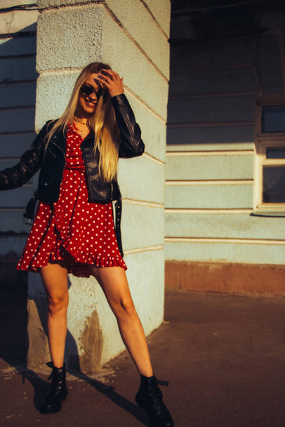 urban street portrait of young beautiful stylish woman in red dress and black leather jacket wearing sunglasses. standing near the old blue wall - Fotoğraf, Görsel
