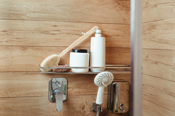 Ein Badeschwamm, eine Körpermassagebürste, ein Penza, eine Flasche Shampoo und Haarbalsam stehen in einem Regal in einem Badezimmer mit Holzwand. Bade- und Duschkonzept - Foto, Bild