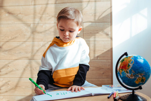 a fair-haired boy is drawing. to get ready for school .there is a globe on the table - Φωτογραφία, εικόνα
