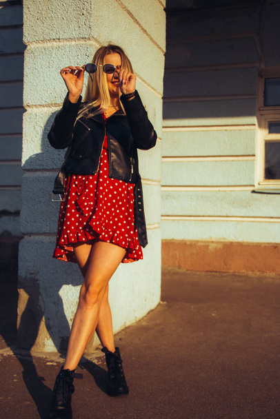 urban street portrait of young beautiful stylish woman in red dress and black leather jacket wearing sunglasses. standing near the old blue wall - Photo, Image