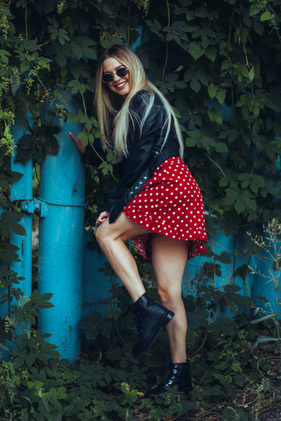 urban street portrait of young beautiful stylish woman in red dress and black leather jacket wearing sunglasses. stands near a blue fence entwined with green plants - Фото, изображение