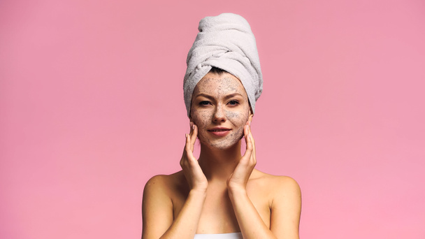 улыбающаяся женщина с махровым полотенцем на голове нанося скраб на лицо изолирован на розовый - Фото, изображение