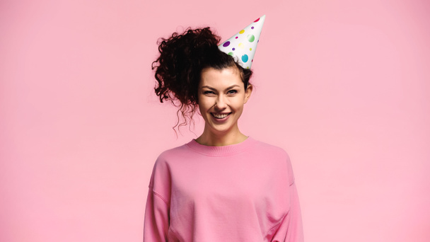 cheerful curly woman in party cap smiling at camera isolated on pink - Fotoğraf, Görsel
