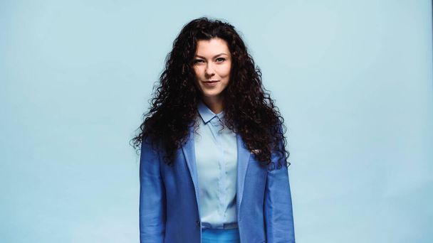positive woman in blue blazer smiling at camera isolated on blue - Foto, afbeelding