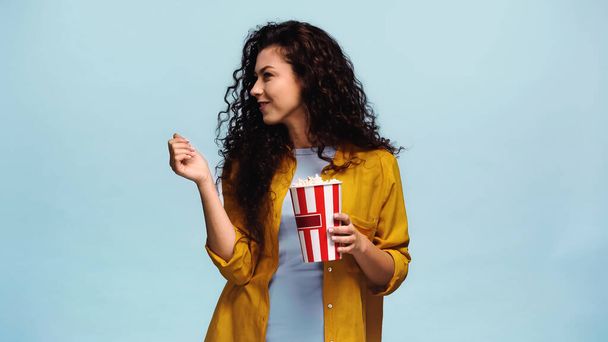 curly woman with popcorn smiling while looking away isolated on blue - Foto, immagini