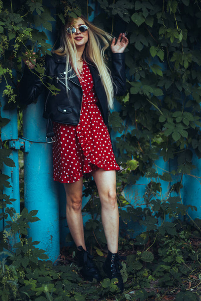 urban street portrait of young beautiful stylish woman in red dress and black leather jacket wearing sunglasses. stands near a blue fence entwined with green plants - Zdjęcie, obraz