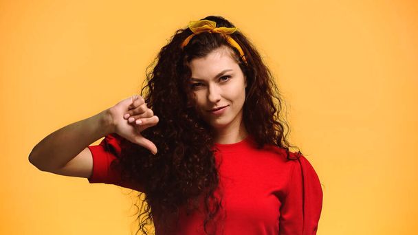 smiling woman in red blouse and headband showing thumb down isolated on orange - Foto, imagen