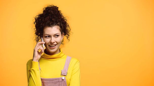 cheerful woman with wavy hair talking on smartphone isolated on orange - Фото, изображение