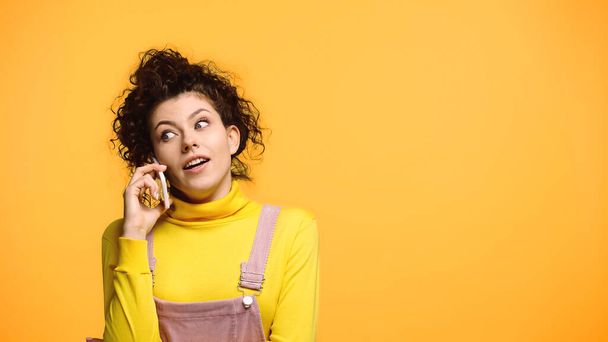 mujer sorprendida en cuello alto amarillo hablando en teléfono inteligente aislado en naranja - Foto, Imagen