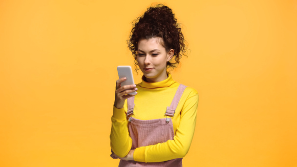 positive woman with curly hair chatting on smartphone isolated on orange - Foto, afbeelding