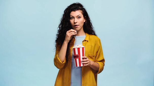 surprised woman in orange shirt eating popcorn isolated on blue - Foto, imagen