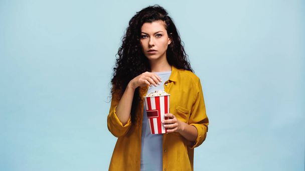 концентрированная женщина держит ведро попкорна изолированы на синий - Фото, изображение