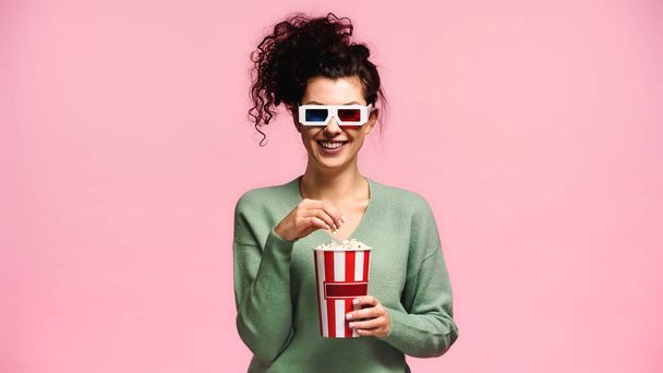 vidám nő zöld pulóver és 3D-s szemüveg eszik popcorn elszigetelt rózsaszín - Fotó, kép
