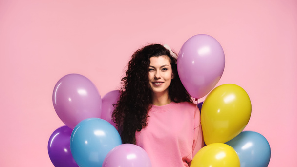 positive brunette woman looking at camera near colorful balloons isolated on pink - 写真・画像