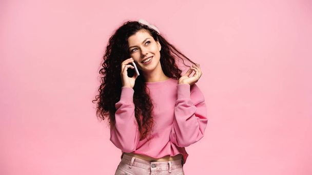 joyful woman touching hair and looking away while talking on smartphone isolated on pink - Photo, Image