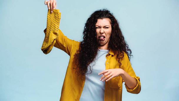 curly woman grimacing and sticking out tongue while holding smelly socks isolated on blue - Foto, afbeelding