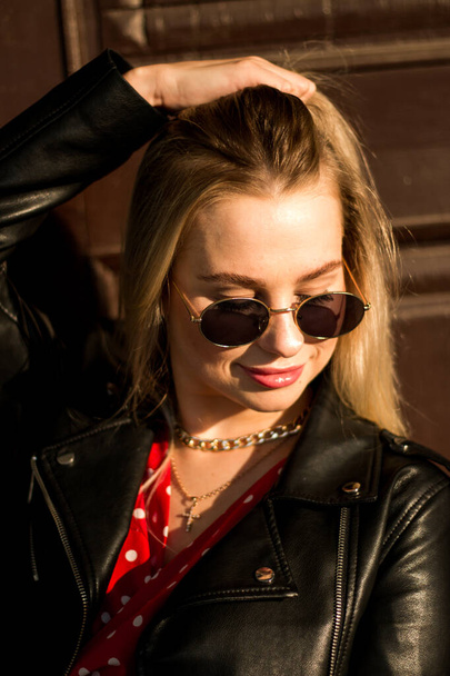 urban street portrait of young beautiful stylish woman in red dress and black leather jacket wearing black sunglasses. Standing near an old brown door, natural evening sunset light - Foto, afbeelding